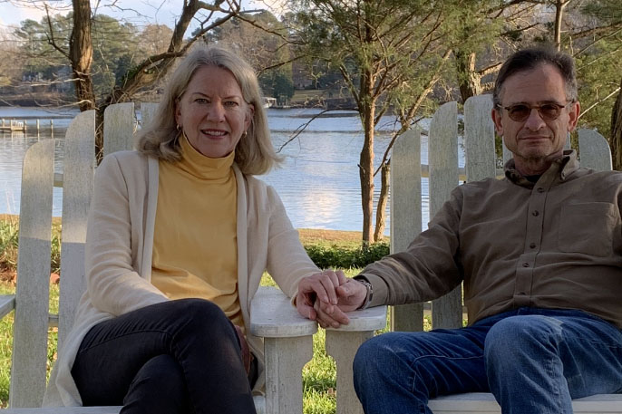Jarva Weiss, a white woman with medium length silver hair, sits on a white Adirondack chair. There is a lake and a smattering of trees behind her. On the right is her husband, Daniel Weiss. He has short, dark hair with silver in it as well. The couple are holding hands. 