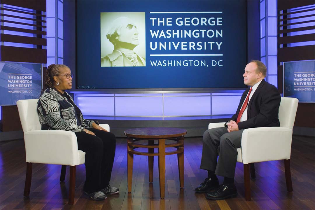 Sherry Molock and Paul Wahlbeck seated on a stage with a George Washington University logo behind them
