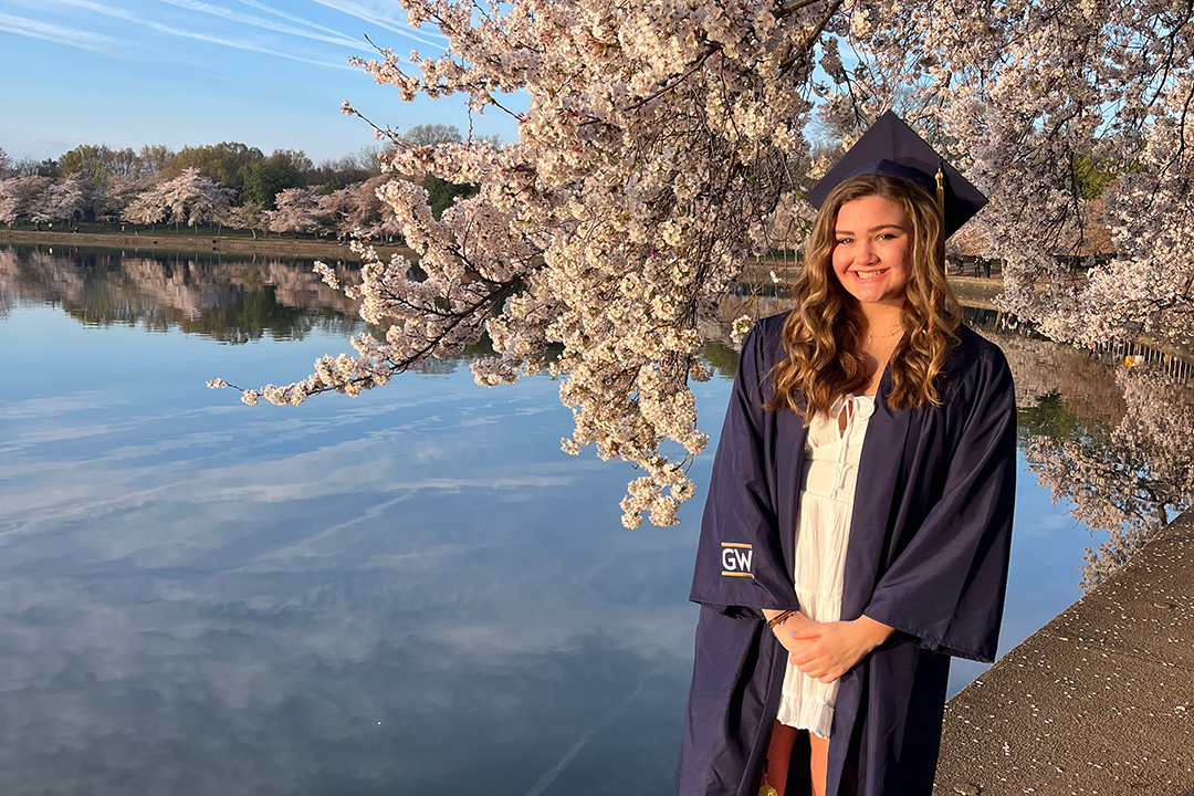 Maggie Connolly wearing graduation robes and gown standing by cherry blossom trees at the Tidal Basin