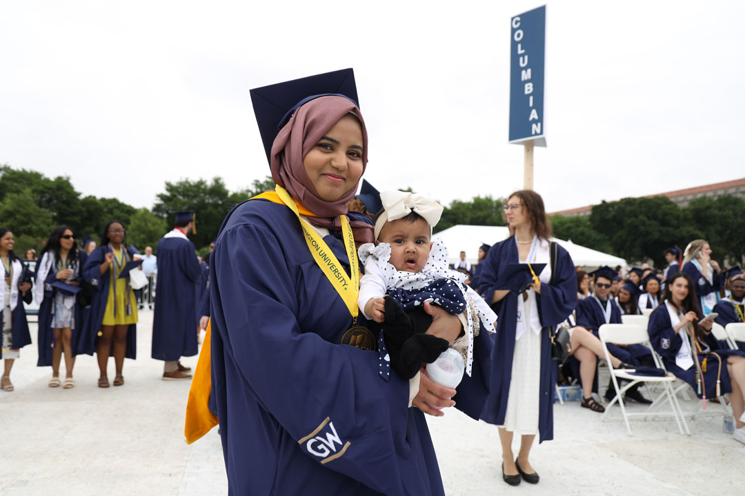 A graduate in cap and gown holds a tiny baby during GW Commencement 2024 on the National Mall
