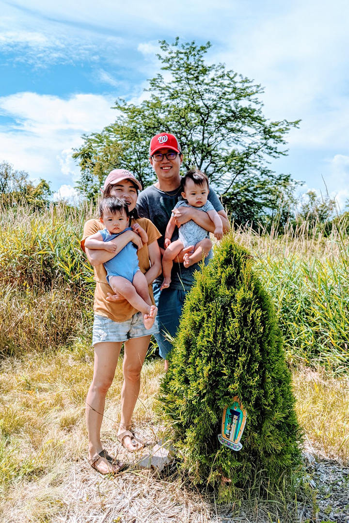 Kao and his wife holding two small children and smiling outdoors in a field with a fir tree