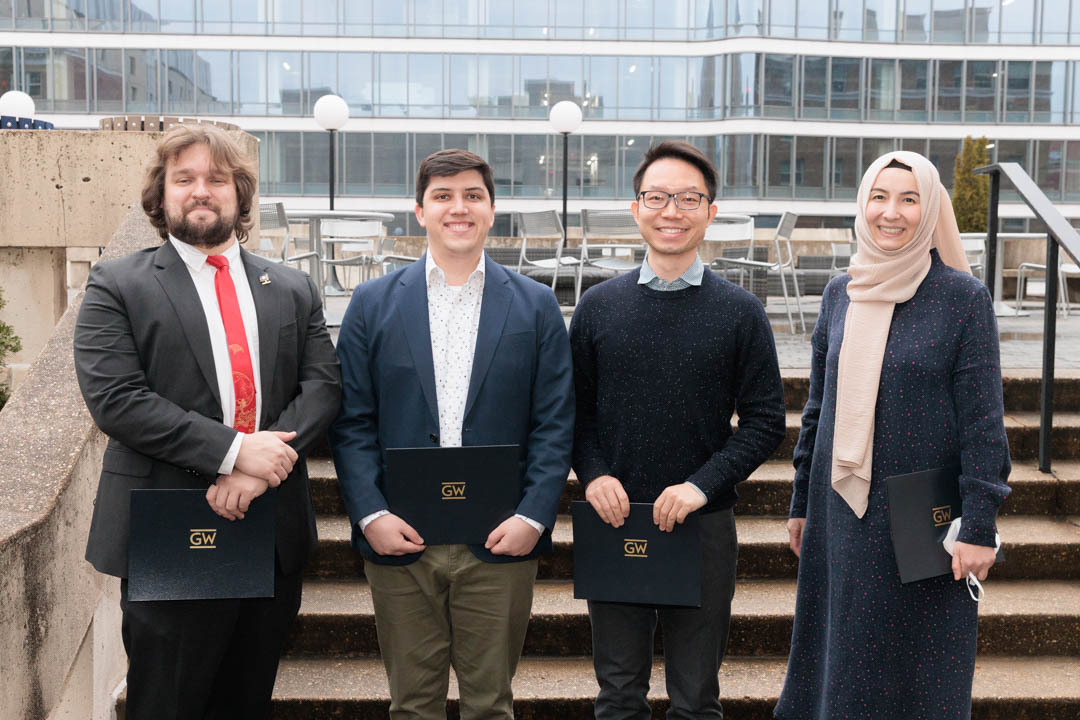 Four doctoral students stand together in front of a GW building smiling