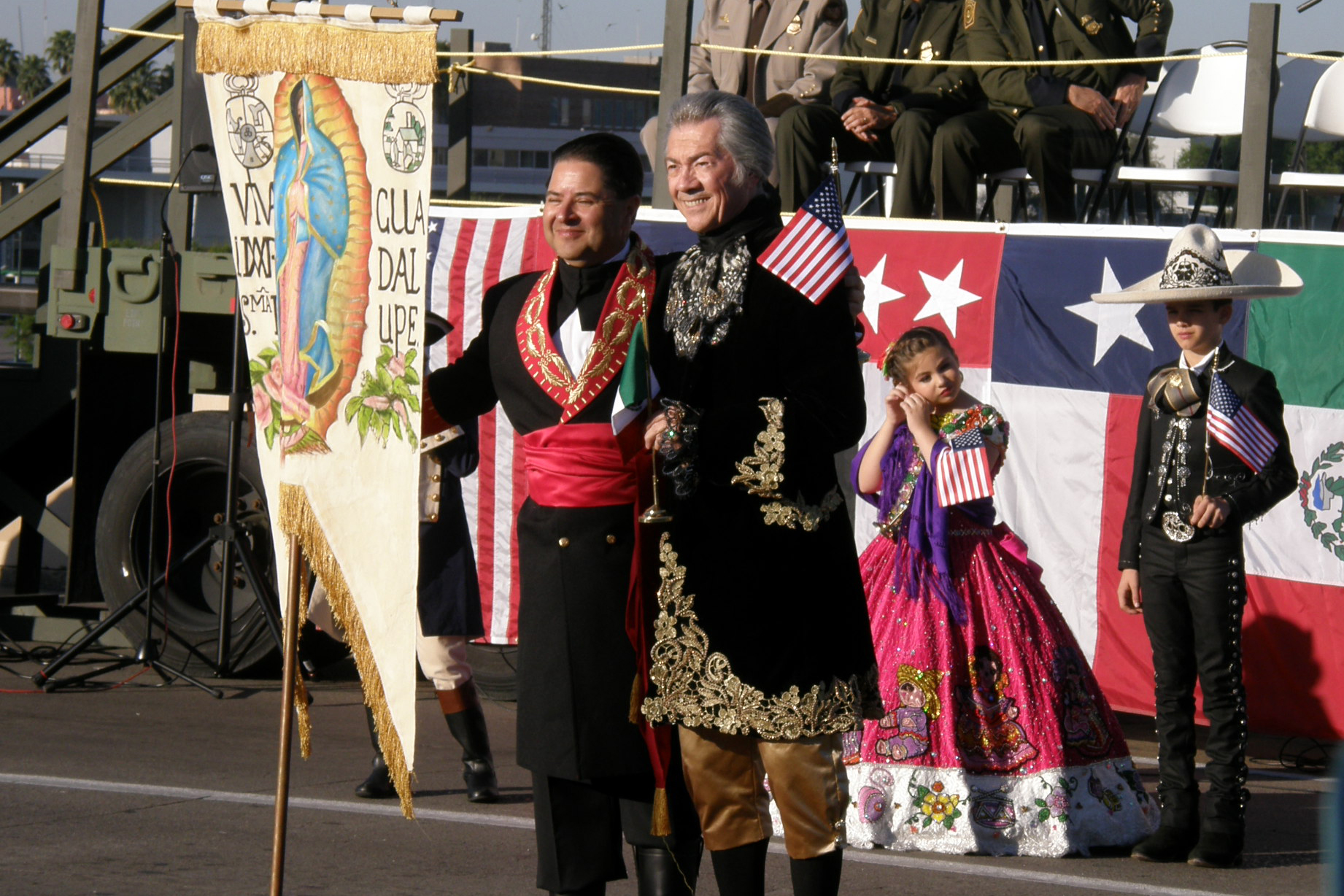 Two officials of the ceremony standing with a banner of the Virgin Mary, two children stand behind them in traditional Mexican festival costumes