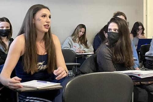 A student on the left sits at a desk in class. She has a GW temporary tattoo on her cheek. Another student sits to the right of her. She has long brown hair and is wearing a black KN95 mask.
