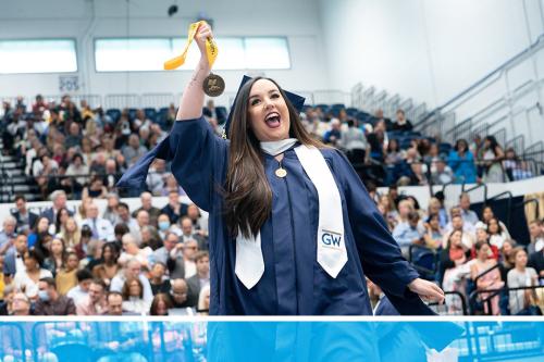 GW student in master's cap and gown holds up her graduation medal in front of a crowded auditorium