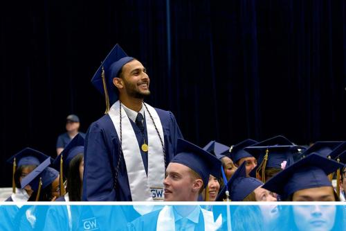 A student in a cap and gown standing and smiling at someone in the crowd at the CCAS 2024 celebration