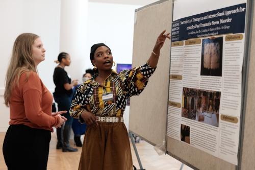 LauraChioma Jones points at her presentation on a wallboard, discussing her research with a fellow student