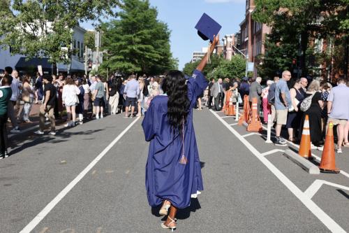 A young woman with her back to the camera wearing a graduation gown and holding her cap in the air