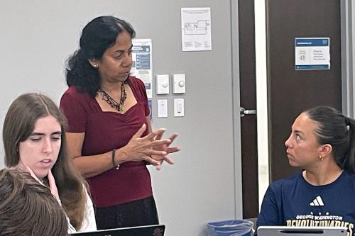 Elizabeth Chacko stands in a classroom gesturing and speaking to two seated students