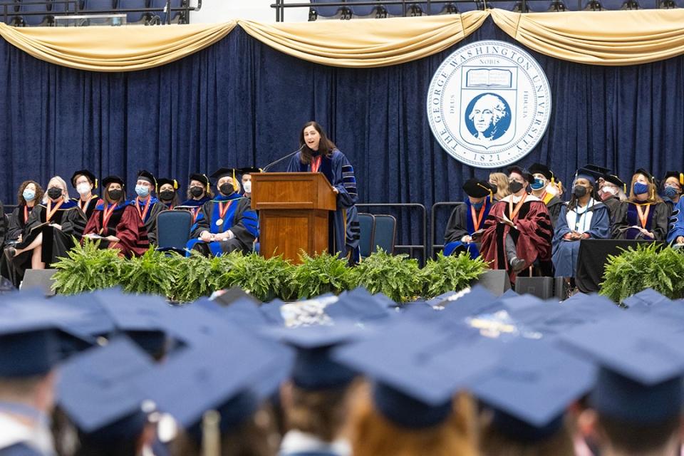 A professor speaking at a GW podium on stage with a sea of graduation caps in the foreground