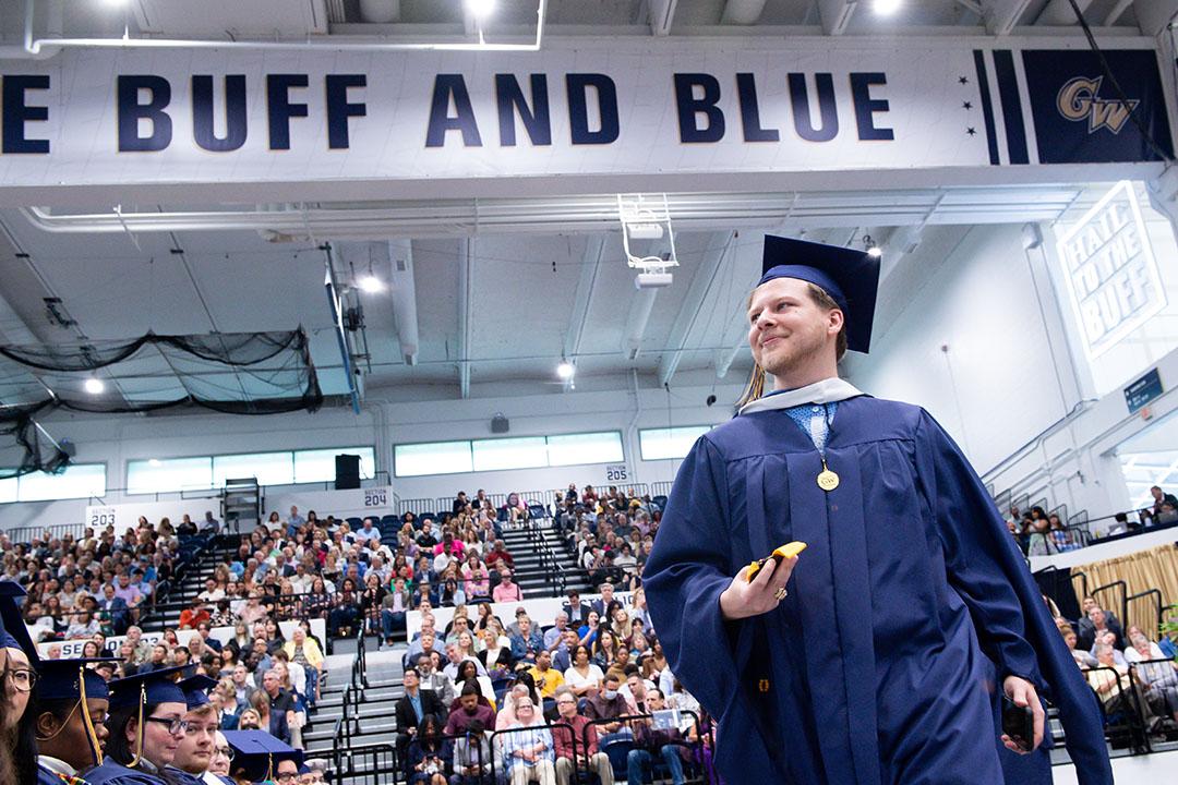 A CCAS master's student in cap and gown underneath the BUFF AND BLUE banner