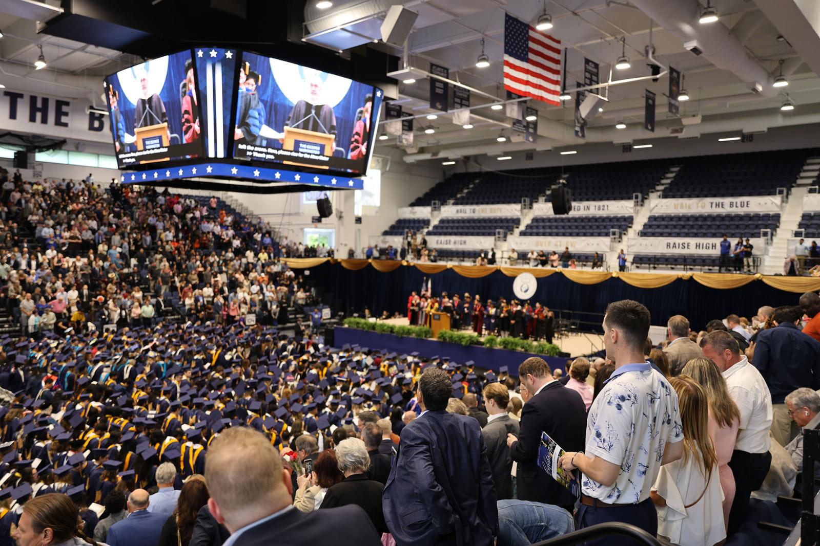 A sea of graduates in a crowd of family and friends at the 2024 celebration for columbian college, with a jumbotron displaying above