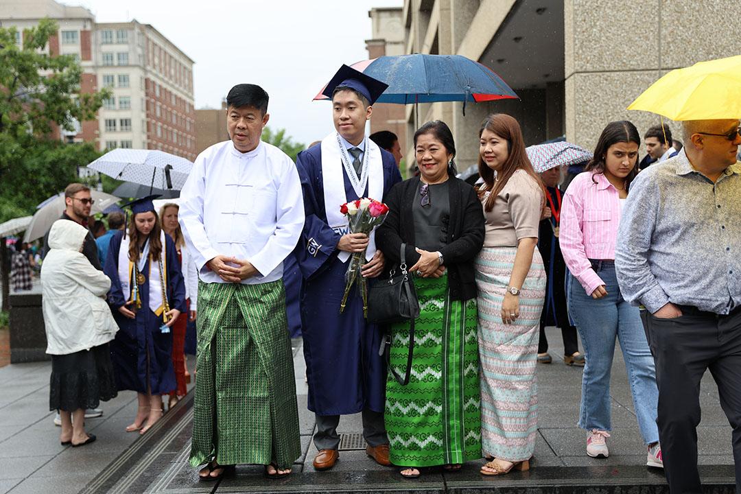 A recent graduate in cap and gown stands outside with his family members on a rainy day