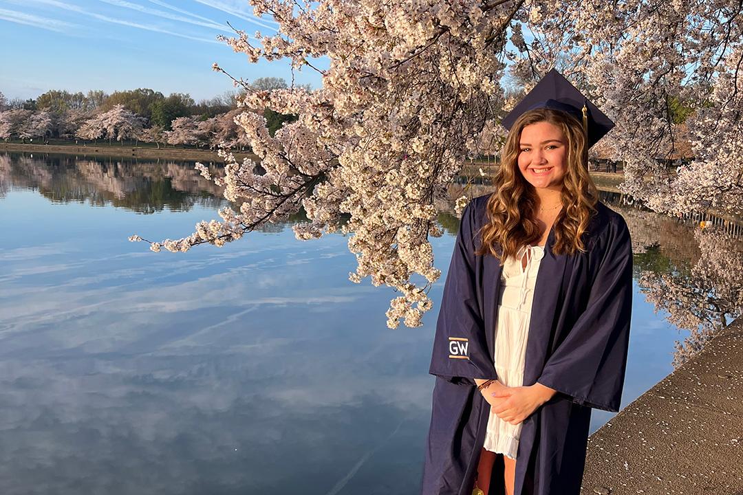 Maggie Connolly wearing graduation robes and gown standing by cherry blossom trees at the Tidal Basin