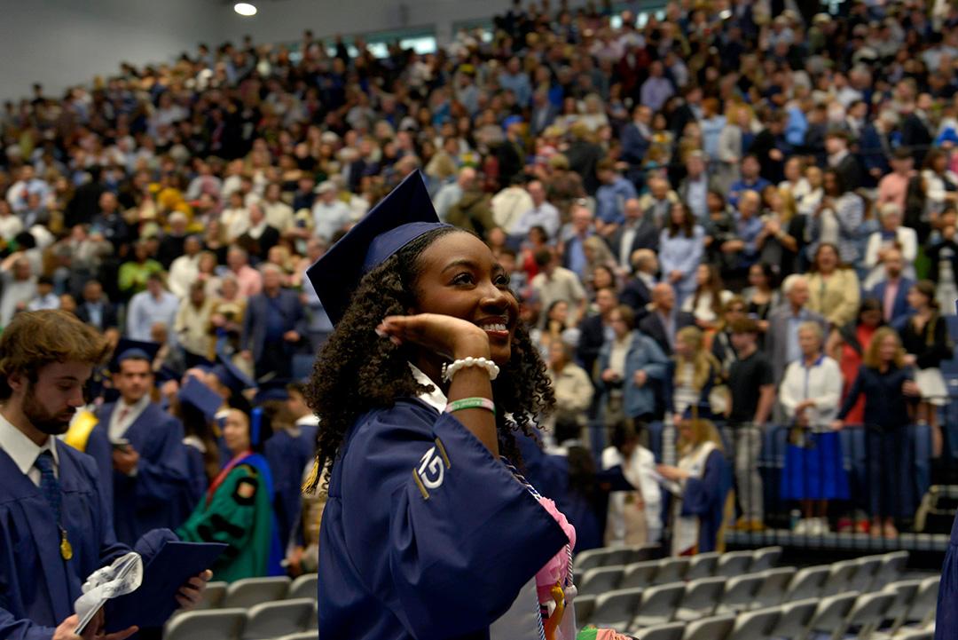 Close up of a student in graduation cap and gown smiling and waving at the audience in the stands