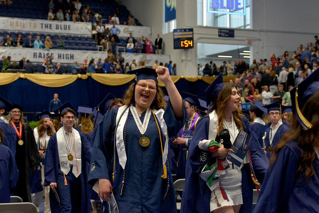An undergraduate student in cap and gown fist bumping and celebrating while processing with fellow graduates