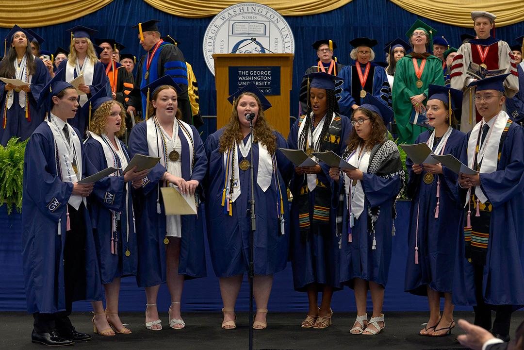 A group of singers in GW graduation regalia standing in front of a stage singing