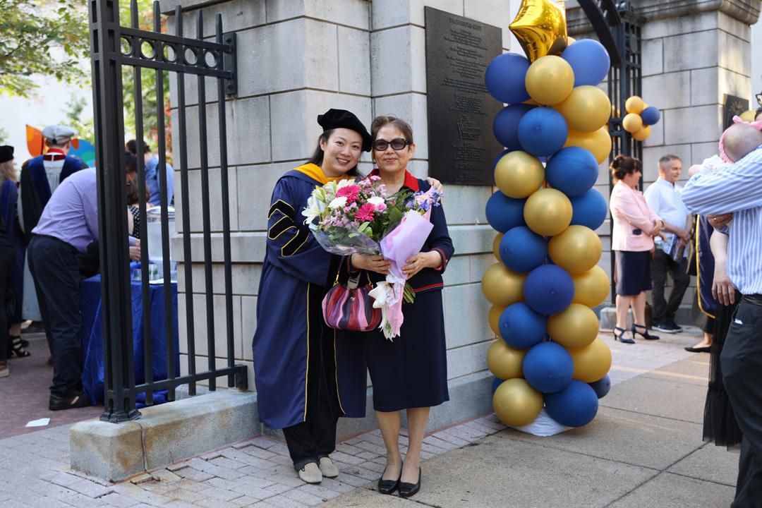 A CCAS doctoral graduate standing next to a woman holding flowers at the 2025 CCAS Doctoral Hooding celebration