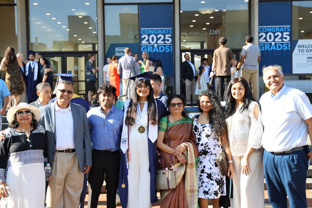 A CCAS GW graduate in cap and gown standing with family and friends on the steps of Gelman Library