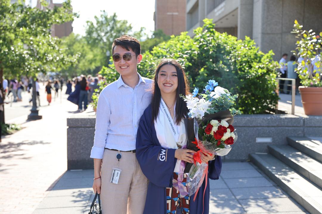 A CCAS graduate in cap and gown holding flowers and standing next to a family member