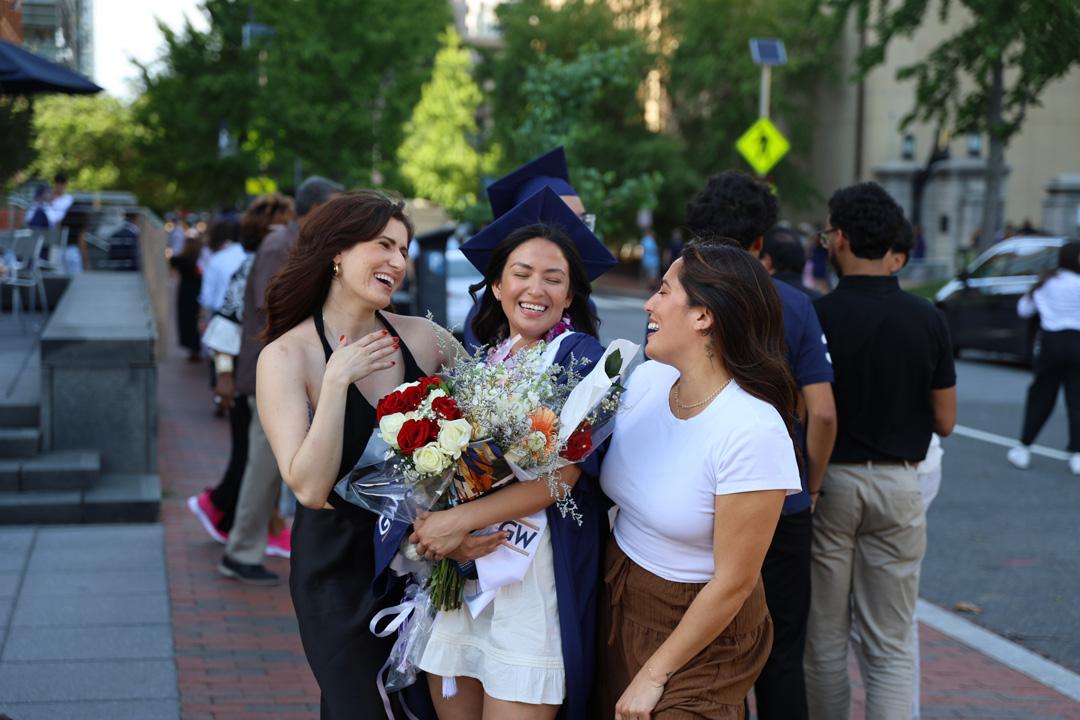A GW undergraduate CCAS student wearing a graduation cap and gown and holding flowers next to her two friends at the 2025 CCAS ceremony