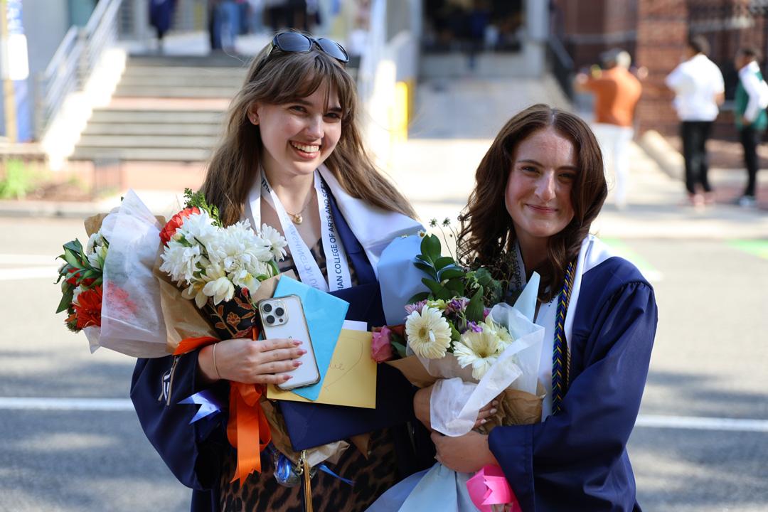 Two CCAS undergraduate students in caps and graduation gowns holding flowers and smiling