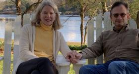 Jarva Weiss, a white woman with medium length silver hair, sits on a white Adirondack chair. There is a lake and a smattering of trees behind her. On the right is her husband, Daniel Weiss. He has short, dark hair with silver in it as well. The couple are holding hands. 